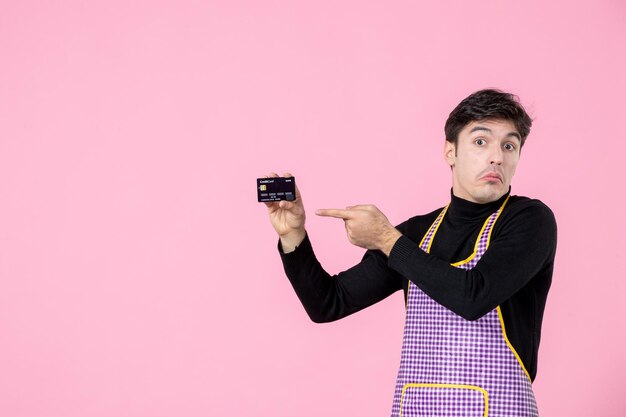 Vista frontal hombre joven en capa con tarjeta bancaria sobre fondo rosa trabajador horizontal colores uniforme cocina profesión jefe trabajo