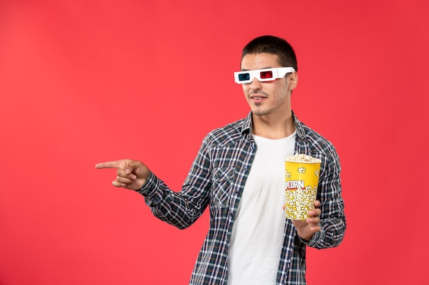 Vista frontal del hombre joven en d gafas de sol con paquete de palomitas de maíz en la pared de color rojo claro películas masculinas cine cine cine