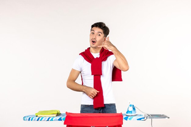 Vista frontal del hombre joven detrás de la tabla de planchar pidiendo llamar sobre fondo blanco hombre hogar esfuerzo limpio emoción quehaceres domésticos ropa de moda