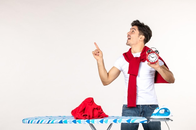 Vista frontal del hombre joven detrás de la tabla de planchar sosteniendo relojes sobre fondo blanco hombre de lavandería a domicilio quehaceres domésticos limpiar emoción tiempo trabajo de color