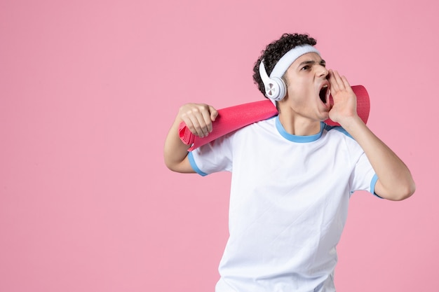 Vista frontal del hombre joven gritando en ropa deportiva con estera de yoga y auriculares