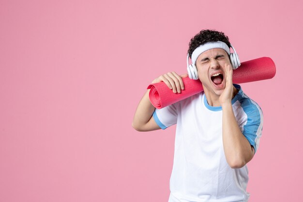 Vista frontal del hombre joven gritando en ropa deportiva con estera de yoga