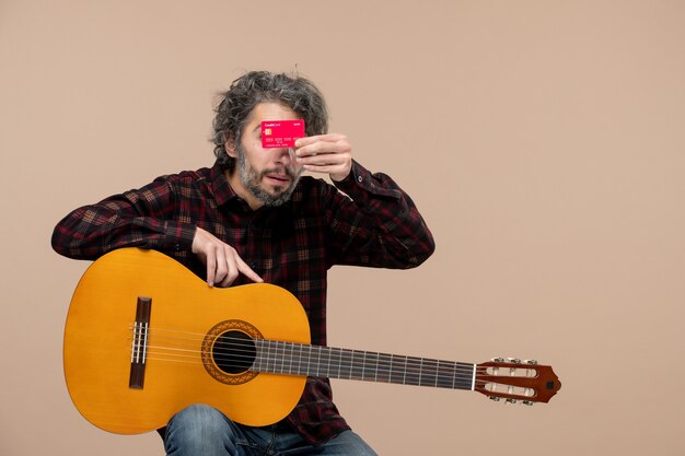Vista frontal del hombre joven con guitarra sosteniendo una tarjeta bancaria roja en la pared rosa