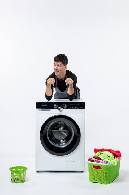 Vista frontal del hombre joven con lavadora y ropa sucia en la pared blanca