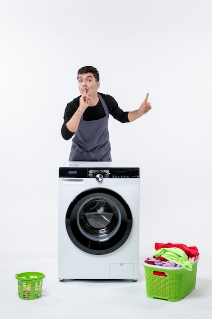 Vista frontal del hombre joven con lavadora y ropa sucia en la pared blanca