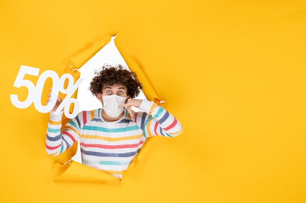 Vista frontal del hombre joven en máscara sosteniendo la escritura sobre el virus amarillo covid de compras- fotos venta pandemia de salud de color