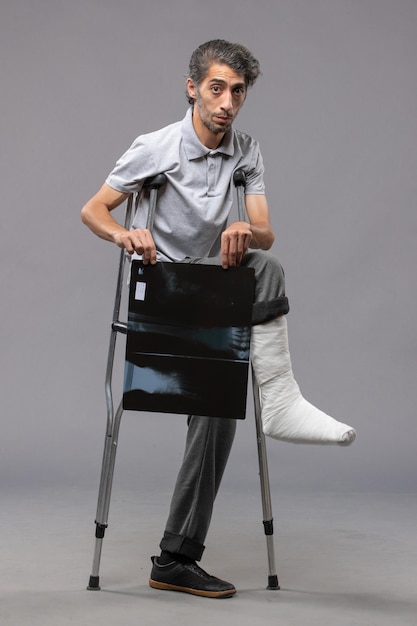 Vista frontal hombre joven con pie roto con muletas y sosteniendo su radiografía en la pared gris dolor deshabilitar pierna rota accidente pie
