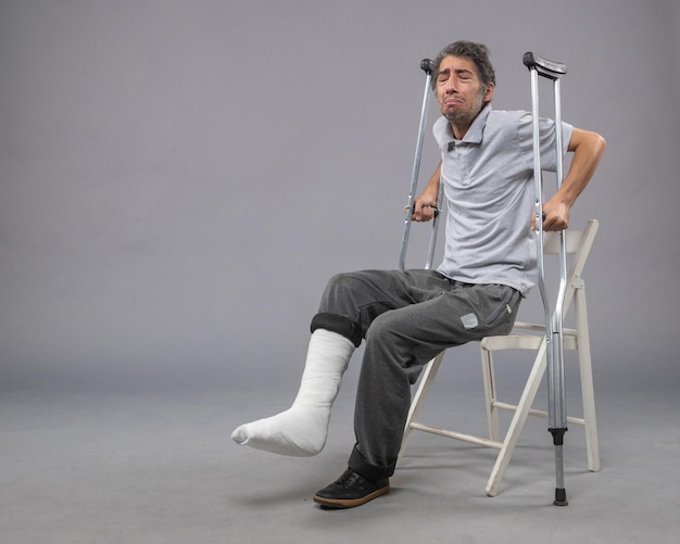 Foto gratuita vista frontal del hombre joven con el pie roto tratando de ponerse de pie sosteniendo muletas en la pared gris pie roto dolor torcer accidente pierna