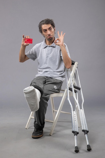 Vista frontal de un hombre joven con un pie roto y un vendaje sosteniendo una tarjeta bancaria en el escritorio gris, el dolor de la pierna torcida, el pie masculino humano
