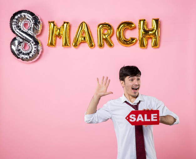 Vista frontal hombre joven con placa de identificación de venta roja con decoración de marzo sobre fondo rosa color vacaciones femenino compras igualdad dinero hombre presente