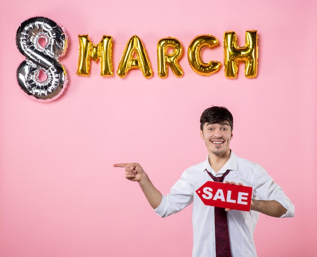 Vista frontal hombre joven con placa de identificación de venta roja con decoración de marzo sobre fondo rosa colores fiesta festiva compras femeninas igualdad dinero hombre presente