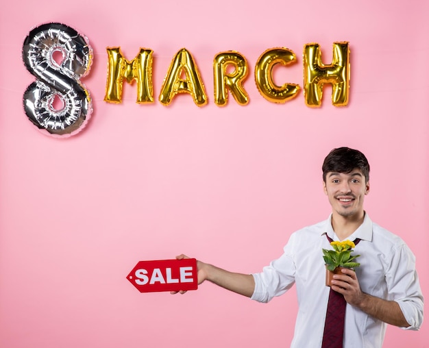 Vista frontal hombre joven con placa de identificación de venta roja con decoración de marzo sobre fondo rosa compras fiesta día de la mujer igualdad dinero hombre color femenino