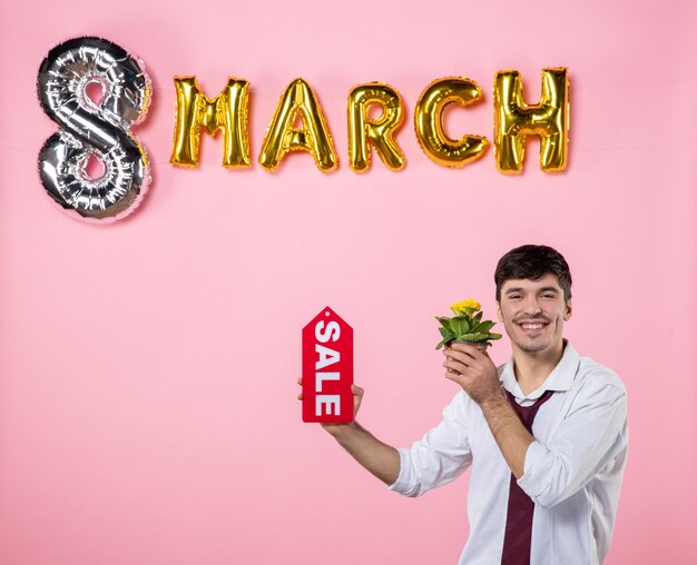 Vista frontal hombre joven con placa de identificación de venta roja con decoración de marzo sobre fondo rosa dinero compras hombre día de la mujer fiesta de la igualdad color de vacaciones