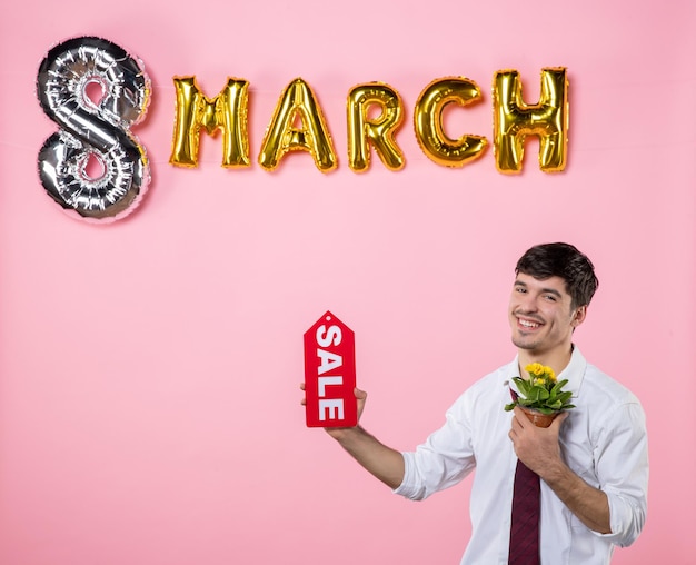 Vista frontal hombre joven con placa de identificación de venta roja con decoración de marzo sobre fondo rosa dinero compras hombre mujer día igualdad femenino vacaciones color