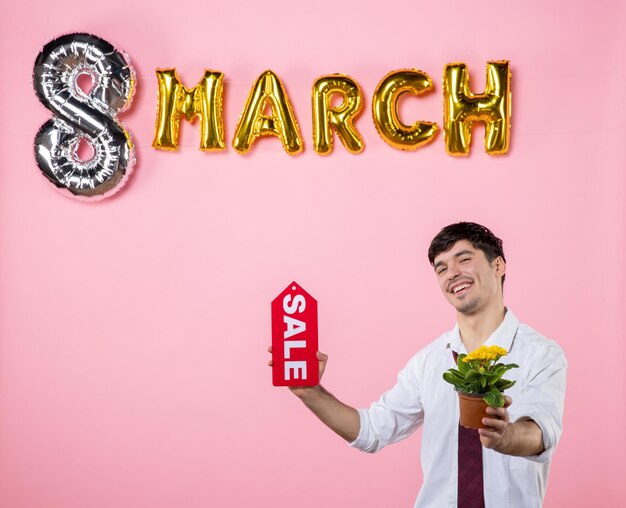 Vista frontal hombre joven con placa de identificación de venta roja con decoración de marzo sobre fondo rosa dinero hombre día de la mujer igualdad fiesta femenina color de vacaciones