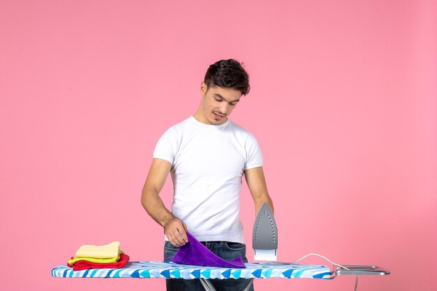 Vista frontal del hombre joven planchando toallas limpias sobre fondo rosa esfuerzo limpio quehaceres domésticos hombre ropa de trabajo a casa