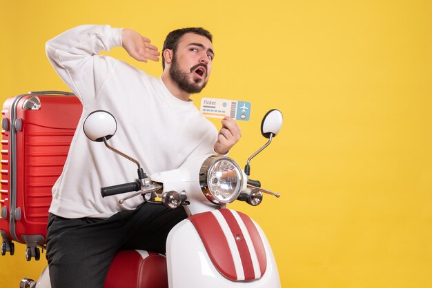 Vista frontal del hombre joven que viaja sentado en la motocicleta con la maleta sujetando el boleto escuchando los últimos chismes sobre fondo amarillo aislado