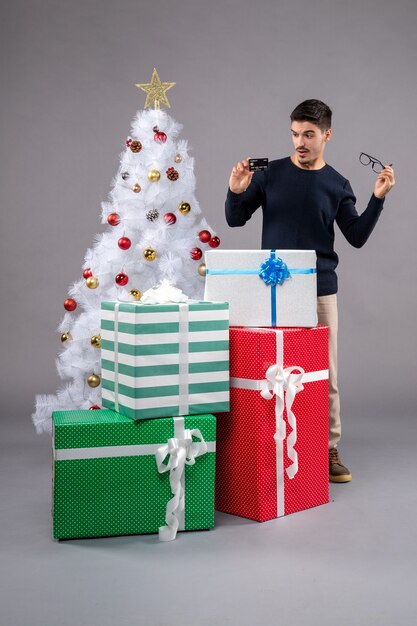 Vista frontal del hombre joven con regalos y tarjeta bancaria en el escritorio gris año nuevo navidad