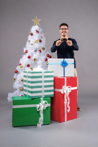 Vista frontal del hombre joven con regalos y tarjeta bancaria en el gris