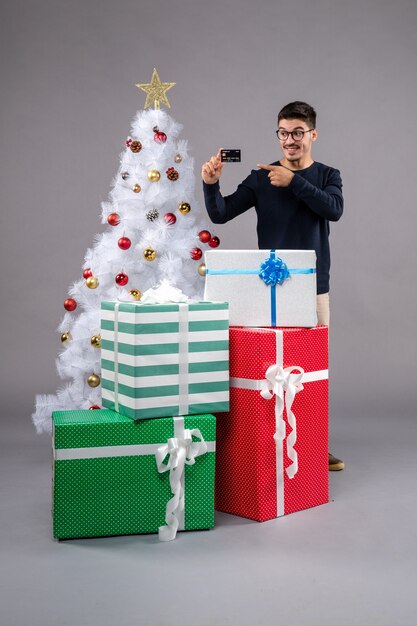 Vista frontal del hombre joven con regalos y tarjeta bancaria en el gris