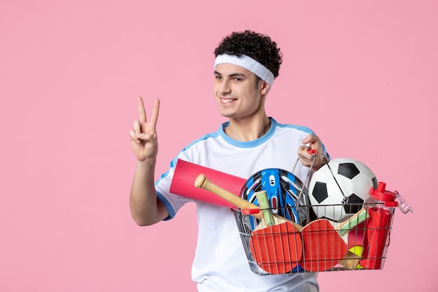 Vista frontal hombre joven en ropa deportiva con canasta llena de cosas deportivas pared rosa