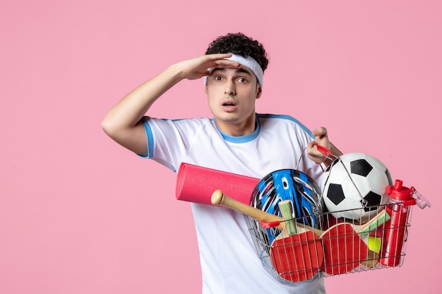 Vista frontal hombre joven en ropa deportiva con canasta llena de cosas deportivas pared rosa