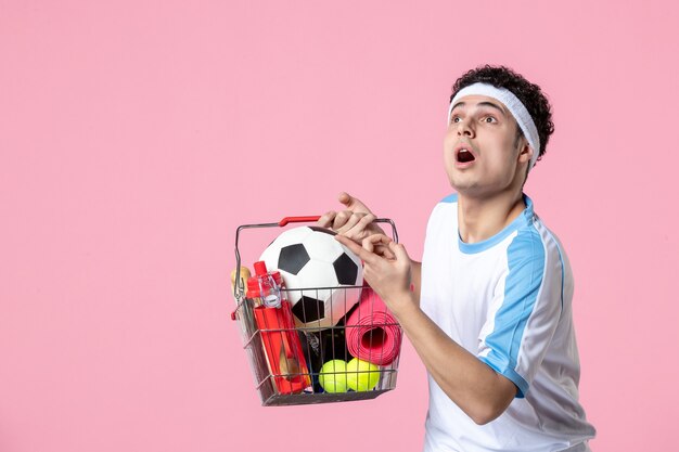 Vista frontal hombre joven en ropa deportiva con canasta llena de cosas deportivas pared rosa