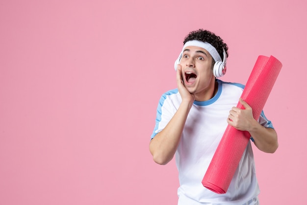 Vista frontal del hombre joven en ropa deportiva con estera de yoga en la pared rosa