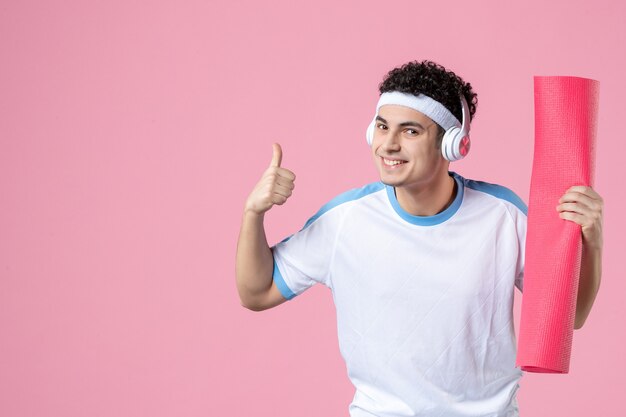 Vista frontal del hombre joven en ropa deportiva con estera de yoga en la pared rosa