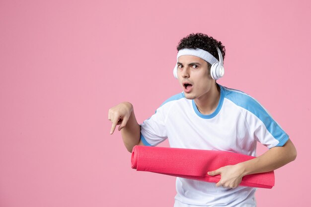 Vista frontal del hombre joven en ropa deportiva con estera de yoga en la pared rosa