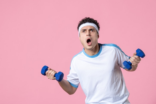 Vista frontal del hombre joven en ropa deportiva trabajando con pesas en la pared rosa