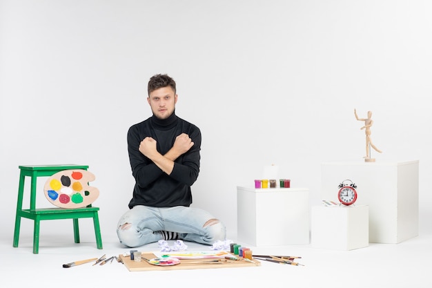 Vista frontal hombre joven sentado alrededor de pinturas y borlas para dibujar en la pared blanca dibujar imagen en color pintura artista pinta arte