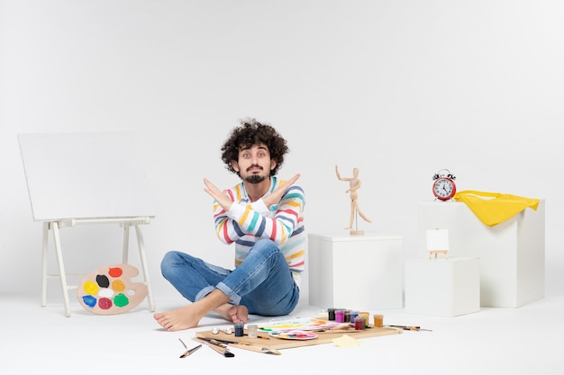 Foto gratuita vista frontal del hombre joven sentado alrededor de pinturas y dibujos en la pared blanca