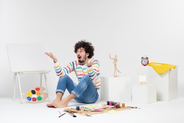 Foto gratuita vista frontal del hombre joven sentado alrededor de pinturas y dibujos en la pared blanca