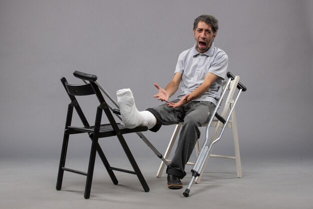 Vista frontal hombre joven sentado con muletas rotas y sufriendo de dolor en la pared gris dolor en el pie accidente piernas rota giro