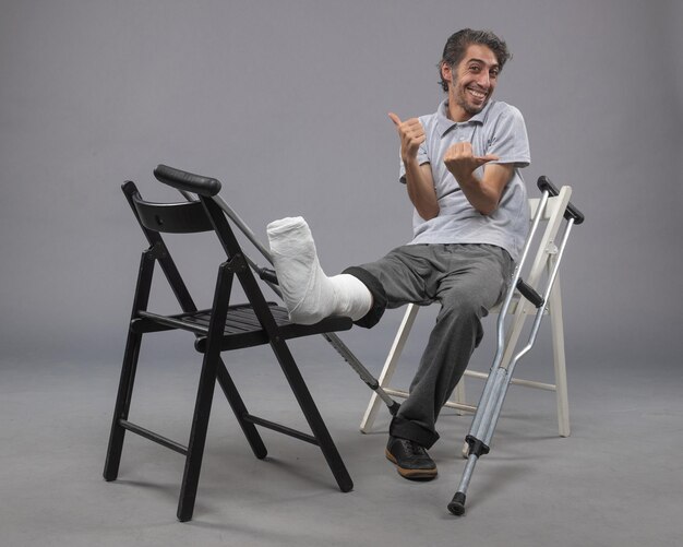 Vista frontal hombre joven sentado con pie roto y muletas en accidentes de pared gris torcer dolor en la pierna del pie roto