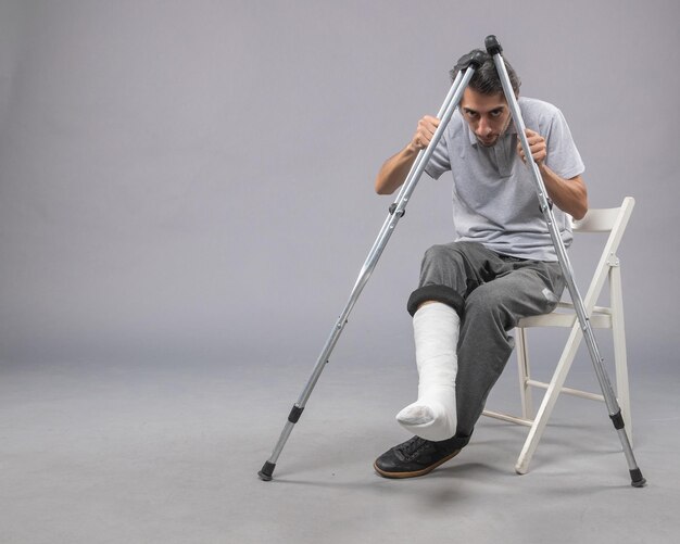 Vista frontal del hombre joven sentado con el pie roto con muletas para caminar sobre el escritorio gris giro de la pierna dolor de accidente pie roto