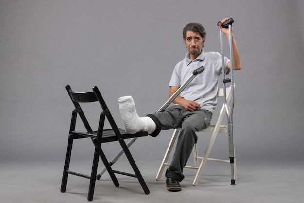 Vista frontal del hombre joven sentado con el pie roto y con muletas en el escritorio gris, giro del pie, dolor roto, accidente, piernas