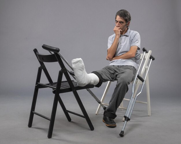 Vista frontal del hombre joven sentado con el pie roto y muletas en el escritorio gris roto accidente piernas del pie dolor torcedura