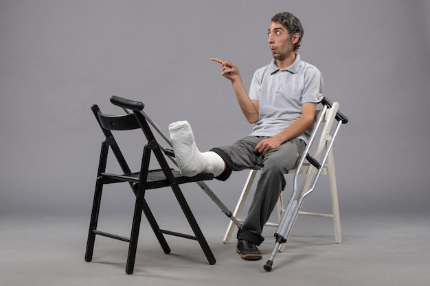 Vista frontal hombre joven sentado con pie roto y muletas en la pared gris accidente pierna rota dolor en el pie torcido