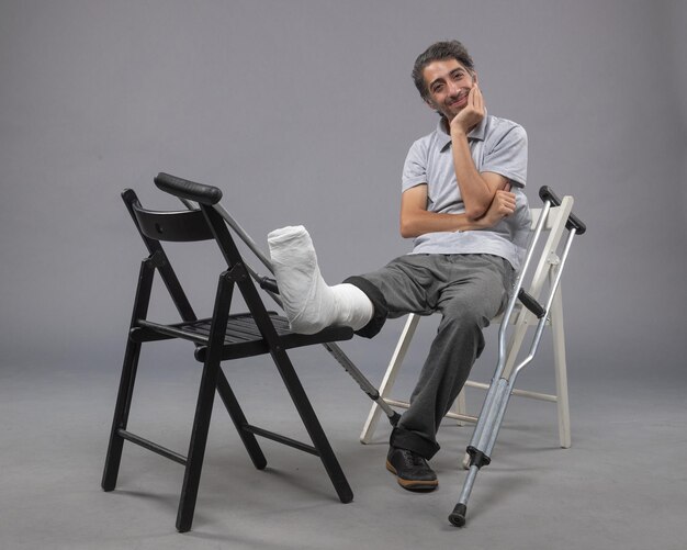 Vista frontal del hombre joven sentado con el pie roto y muletas en la pared gris accidente torcer el dolor de las piernas del pie roto