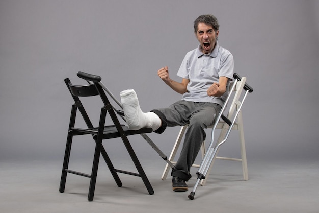 Vista frontal del hombre joven sentado con el pie roto y muletas en la pared gris dolor en el pie accidente pierna rota giro