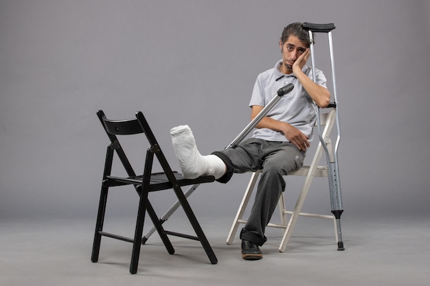 Vista frontal del hombre joven sentado con el pie roto y muletas en la pared gris dolor en el pie accidente piernas rota giro