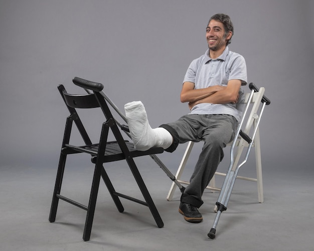 Vista frontal del hombre joven sentado con el pie roto y muletas en la pared gris rota accidente torcedura del pie dolor en las piernas