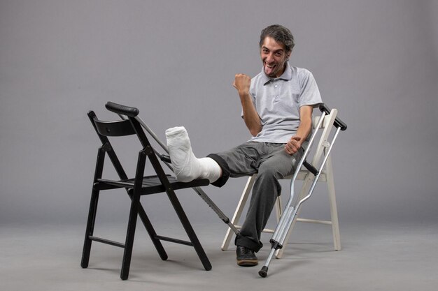 Vista frontal hombre joven sentado con pie roto y muletas regocijándose en la pared gris dolor accidente pierna rota pie torcido