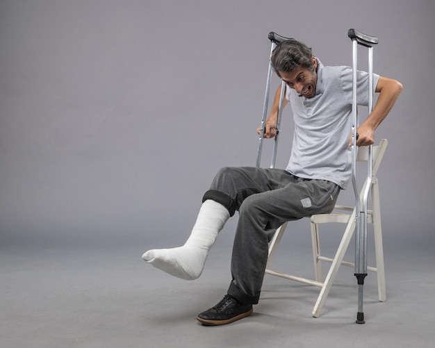 Foto gratuita vista frontal del hombre joven sentado con el pie roto y sosteniendo muletas en la pared gris del pie roto el dolor de la pierna torcer accidente