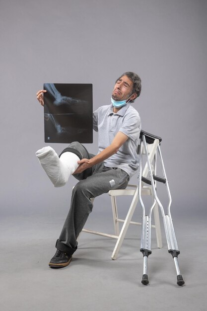 Vista frontal de un hombre joven sentado con un pie roto y sosteniendo una radiografía en el escritorio gris dolor en la pierna accidente torcedura del pie masculino