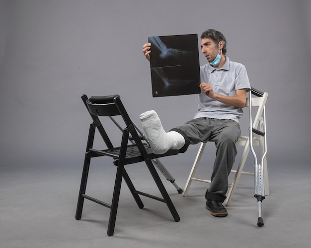 Foto gratuita vista frontal de un hombre joven sentado con un pie roto y sosteniendo una radiografía de la misma en la pared gris.