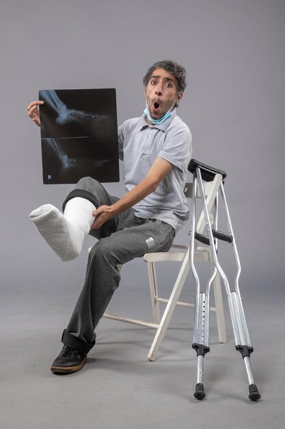 Vista frontal del hombre joven sentado con el pie roto y sosteniendo una radiografía en la pared gris dolor en las piernas accidente torcedura del pie masculino