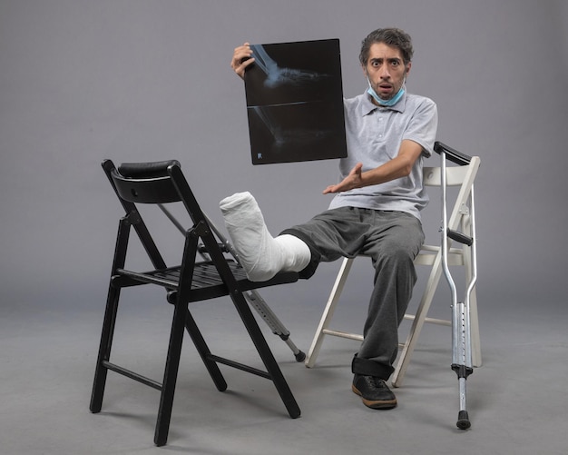 Vista frontal de un hombre joven sentado con un pie roto y sosteniendo una radiografía en la pared gris, dolor de torsión, pierna rota, accidente masculino